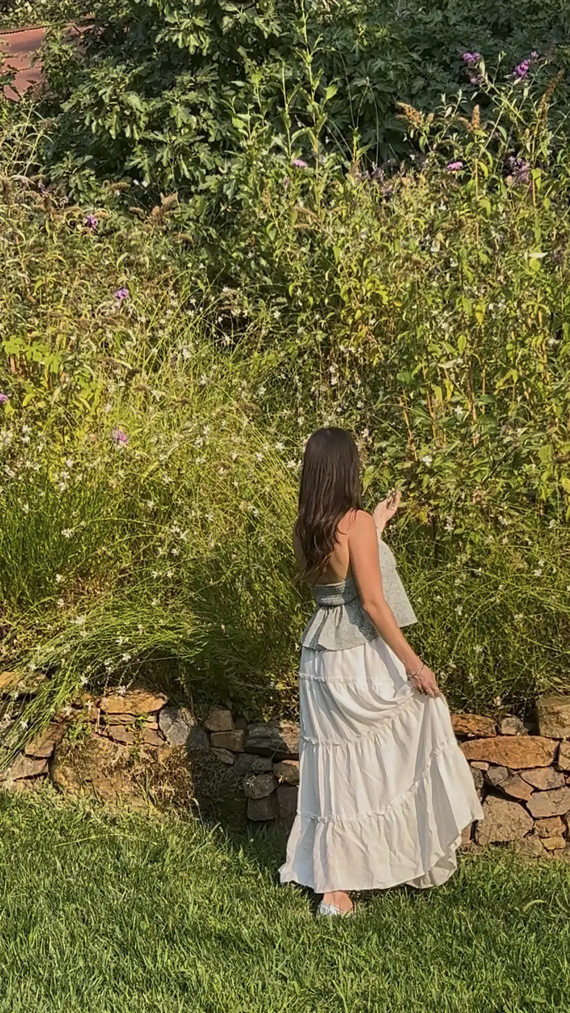 Jenny Hobbs walking a garden path in a long dress, surrounded by tall grasses